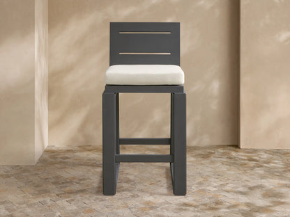 Dark gray Canyon Outdoor Aluminum Barstool with a slatted back and light beige cushion, on a mosaic patio with textured beige walls.
