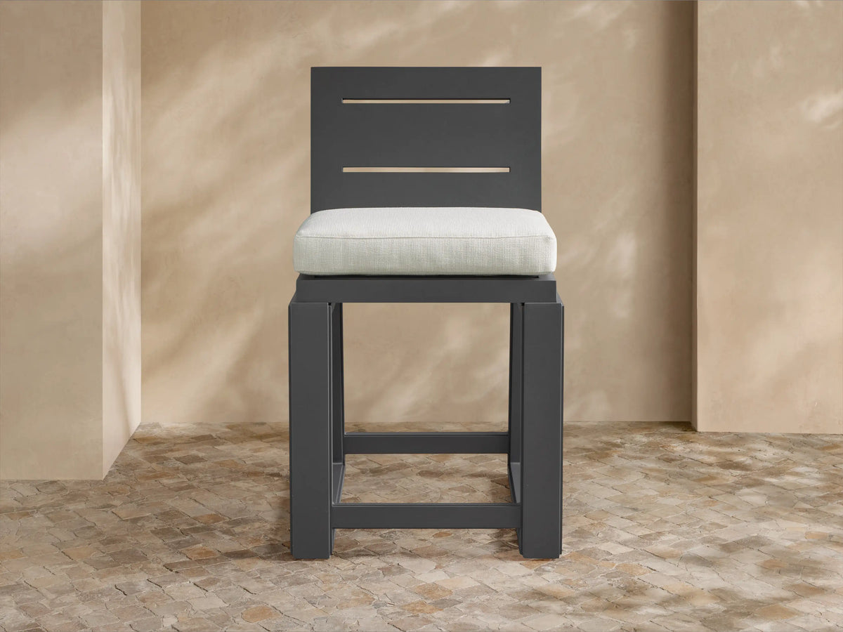Canyon Outdoor Aluminum Counter Stool with charcoal frame, slated back, and light cream cushion on a patio with tiled floor.