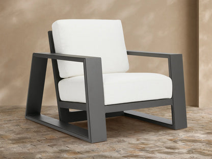Canyon Outdoor Aluminum Lounge Chair with a dark gray angular aluminum frame and thick white cushions, angled on a stone patio.