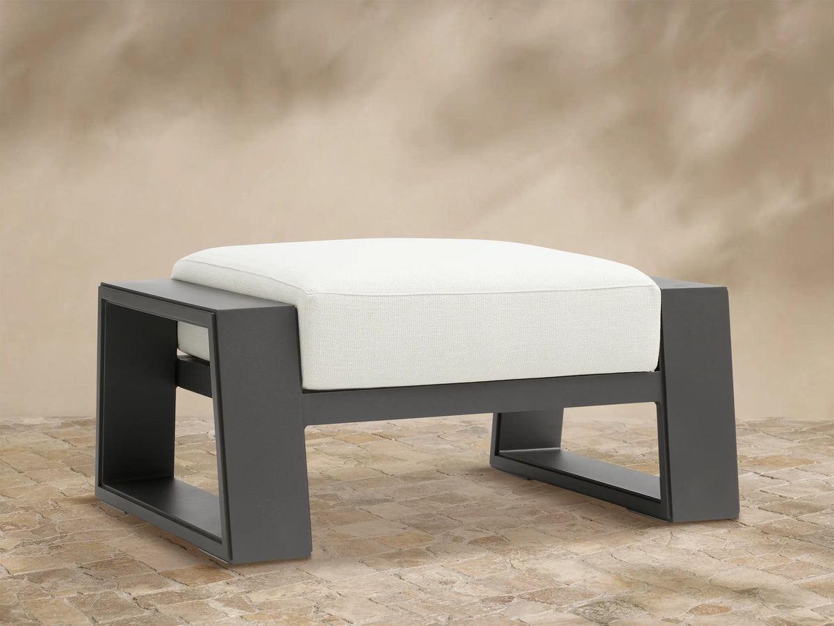 Canyon Outdoor Aluminum Ottoman: a modern, dark gray aluminum frame with a thick, white cushion, on a textured stone patio.