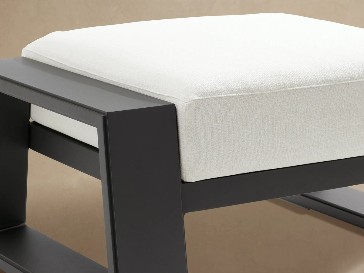 Canyon Outdoor Aluminum Ottoman with a dark gray, modern square frame and a thick, light off-white cushion, on a patio.