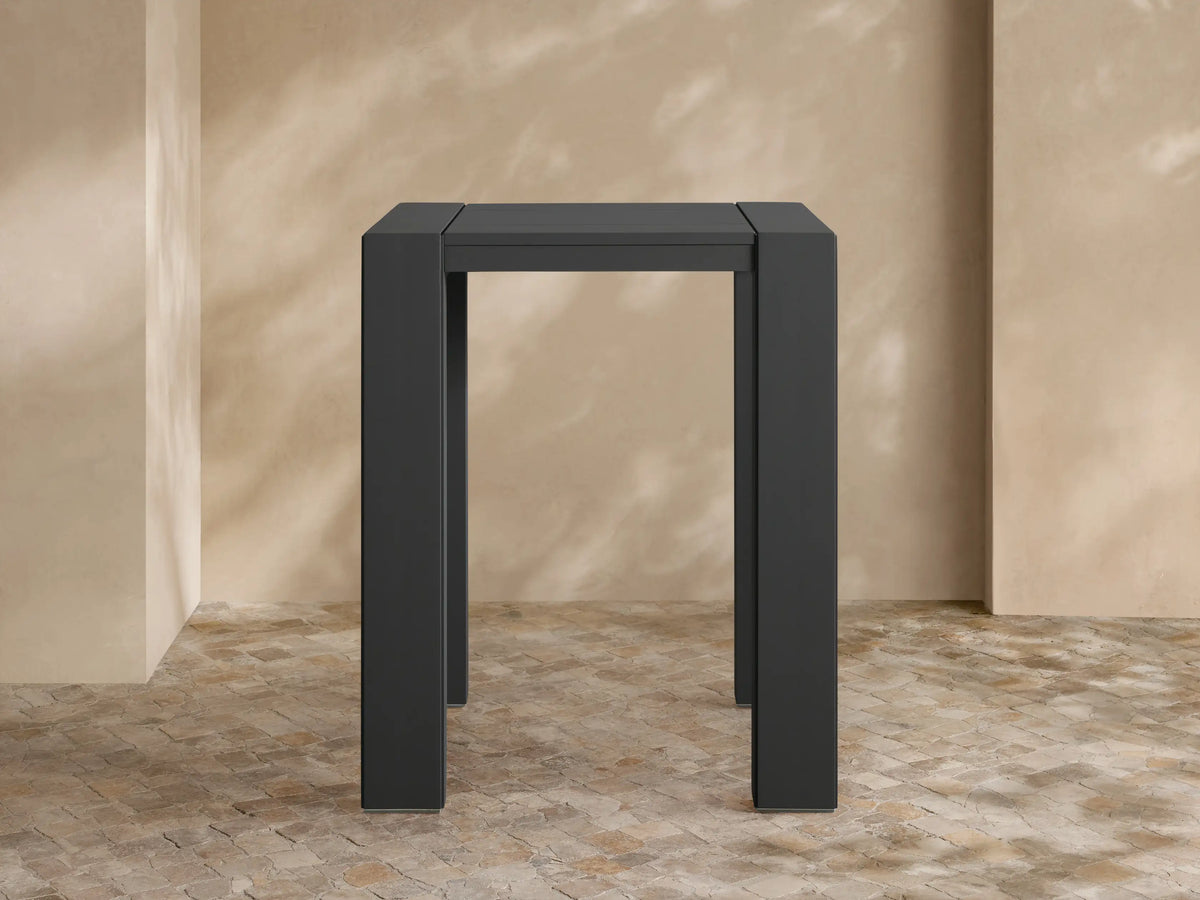 Front view of dark gray Canyon Outdoor Aluminum Bar Table with square top & thick legs, on a stone patio against a textured wall.