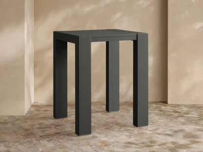 Front view of dark gray Canyon Outdoor Aluminum Bar Table with square top & thick legs, on a stone patio against a textured wall.