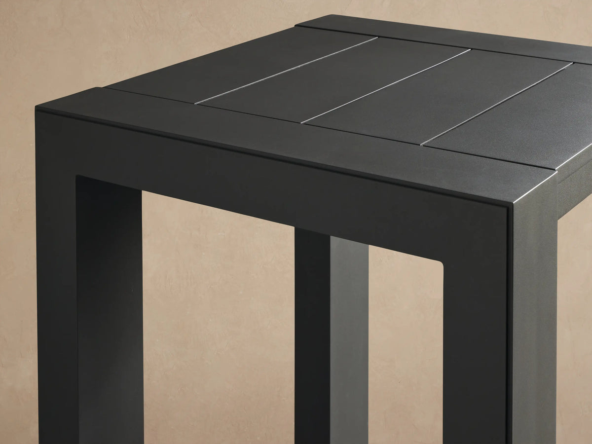 Dark gray square Canyon Outdoor Aluminum Bar Table with slatted top and 4 thick legs on a stone patio.