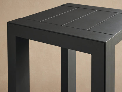 Dark gray square Canyon Outdoor Aluminum Bar Table with slatted top and 4 thick legs on a stone patio.