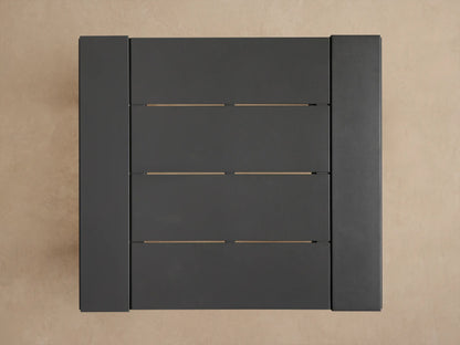 Close-up of a dark gray Canyon Outdoor Aluminum Bar Table. It has a slatted top and thick square legs, against a beige wall.