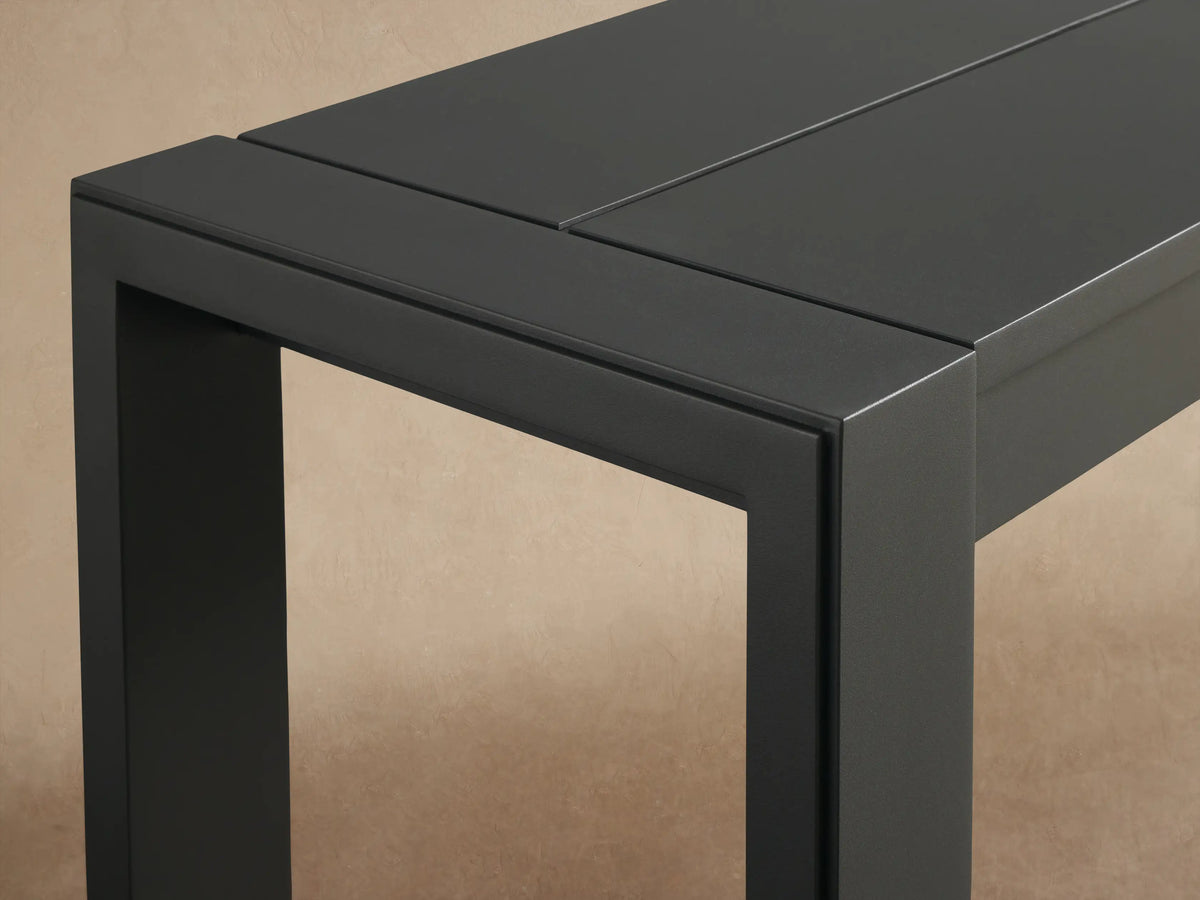 Dark gray Canyon Outdoor Aluminum Dining Bench, sleek design, slatted top, square legs, against a textured beige wall.