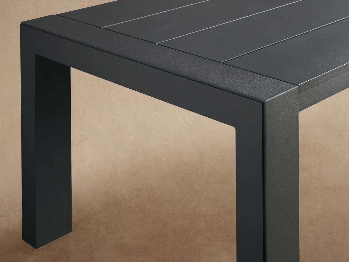 Dark gray Canyon Outdoor Aluminum Dining Table, rectangular with slatted top & U-shaped legs, on a patio with shadow-dappled wall.