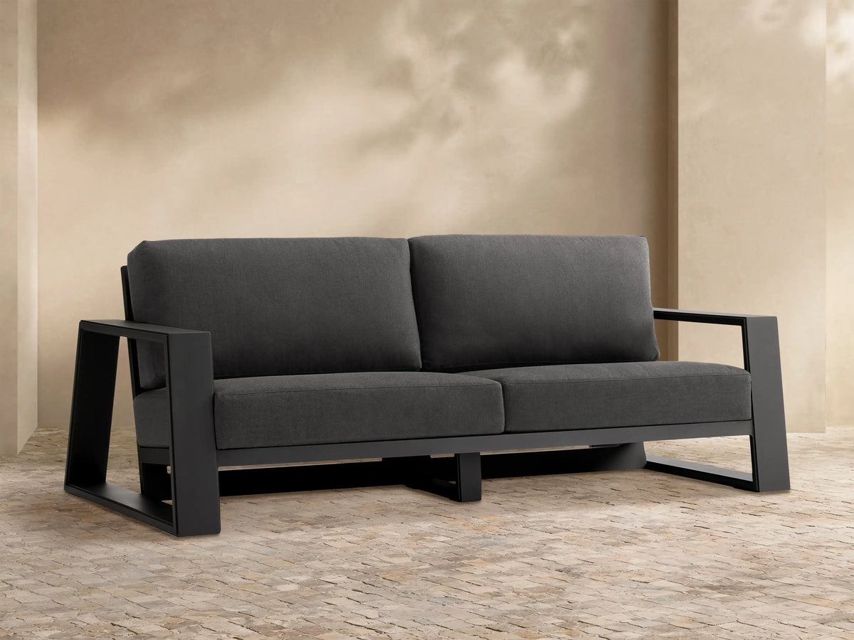 Canyon Outdoor Aluminum Sofa, dark gray cushions on a dark gray aluminum frame, against a beige wall and stone patio.