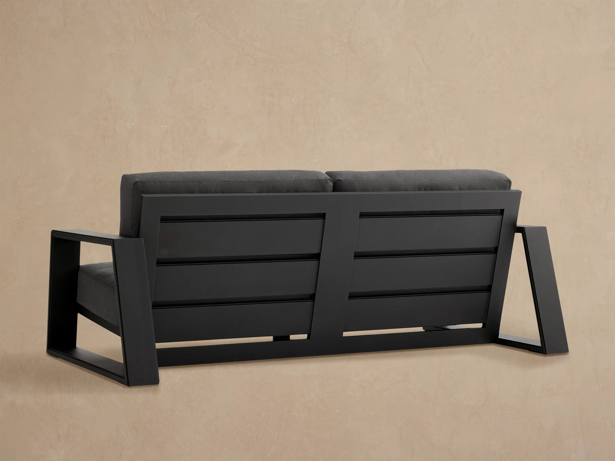 Angled close-up of dark gray Canyon Outdoor Aluminum Sofa with wide armrest, plush cushions, and slatted back against beige wall.