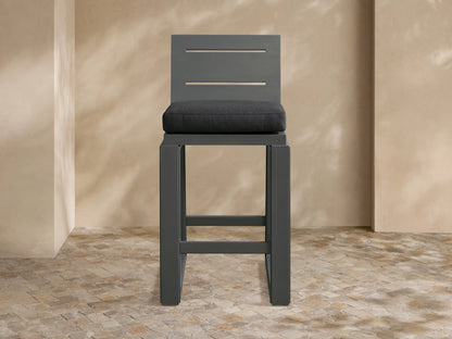 Canyon Outdoor Aluminum Barstool. Dark charcoal aluminum frame, slatted back, and gray fabric cushion, on a patio.