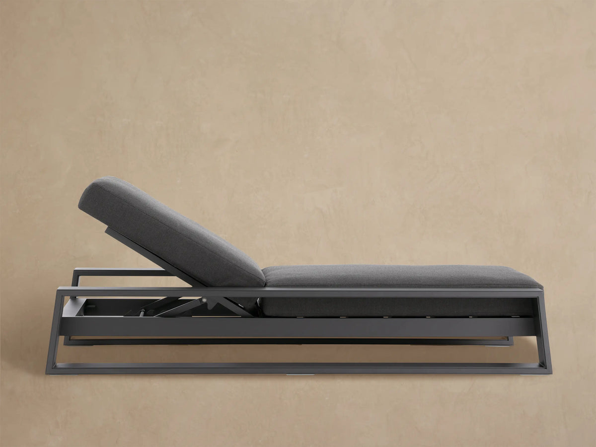 Modern Canyon Outdoor Aluminum Chaise in dark gray with raised backrest, sleek frame, and thick cushions. On a stone patio.