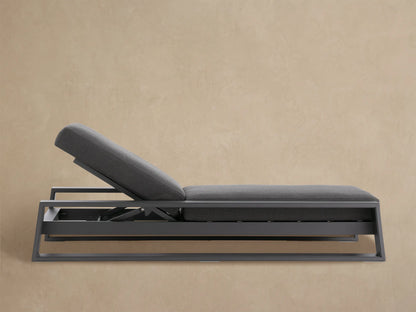 Modern Canyon Outdoor Aluminum Chaise in dark gray with raised backrest, sleek frame, and thick cushions. On a stone patio.