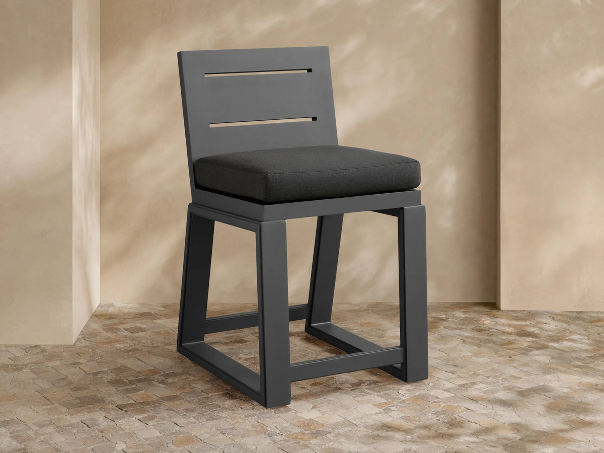 Canyon Outdoor Aluminum Counter Stool, dark gray, with a slatted back and dark cushion, on a textured stone patio.