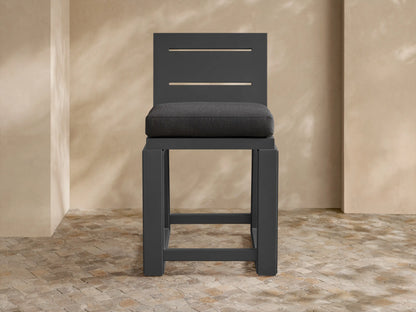 Canyon Outdoor Aluminum Counter Stool, dark gray, with a slatted back and dark cushion, on a textured stone patio.