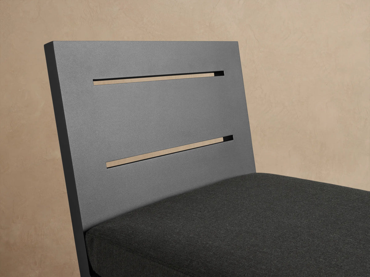 Canyon Outdoor Aluminum Counter Stool in dark gray with a slatted back and cushion, on a stone patio against a beige wall.