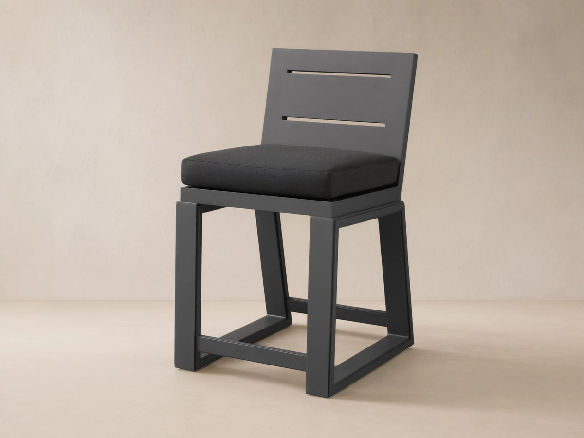 Canyon Outdoor Aluminum Counter Stool, dark gray, with a slatted back and dark cushion, on a textured stone patio.