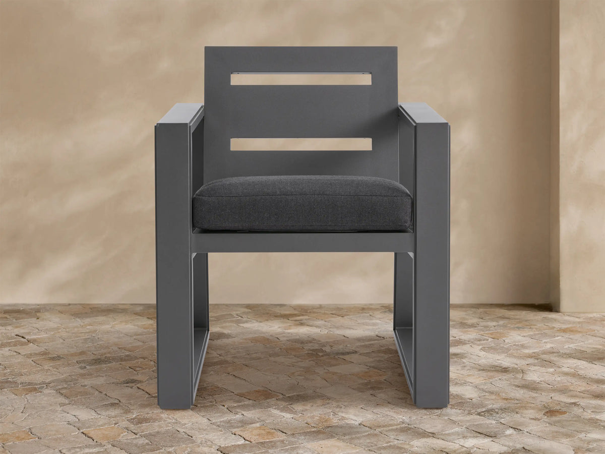 Modern Canyon Outdoor Aluminum Dining Arm Chair, angled, with a gray frame, slatted back, and dark gray cushion on a stone floor.