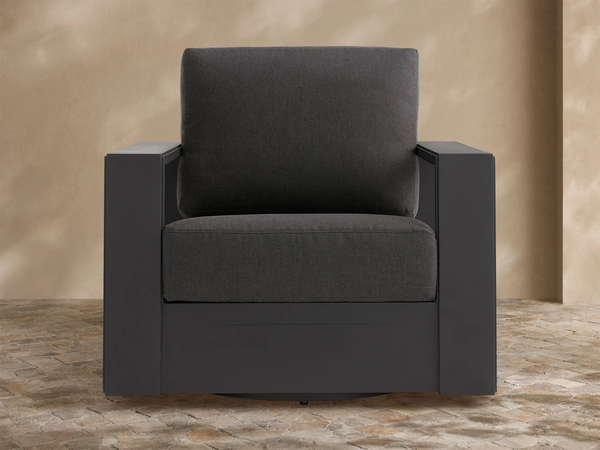 Canyon Outdoor Aluminum Swivel Chair, angled right. Dark charcoal powder-coated frame with thick gray cushions on a stone patio.