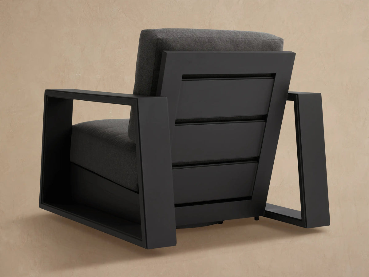 Side view of a modern Canyon Outdoor Swivel Chair. Dark gray aluminum frame and matching cushions against a beige wall.