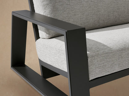 Canyon Outdoor Aluminum Sofa, dark gray aluminum frame, light gray cushions, against a textured beige wall on a patio.