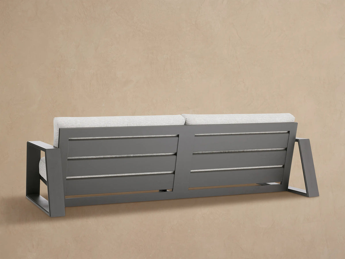 Close-up of Canyon Outdoor Aluminum Sofa's dark gray aluminum armrest & light gray fabric cushions against a tan wall.