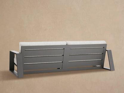 Close-up of Canyon Outdoor Aluminum Sofa's dark gray aluminum armrest & light gray fabric cushions against a tan wall.