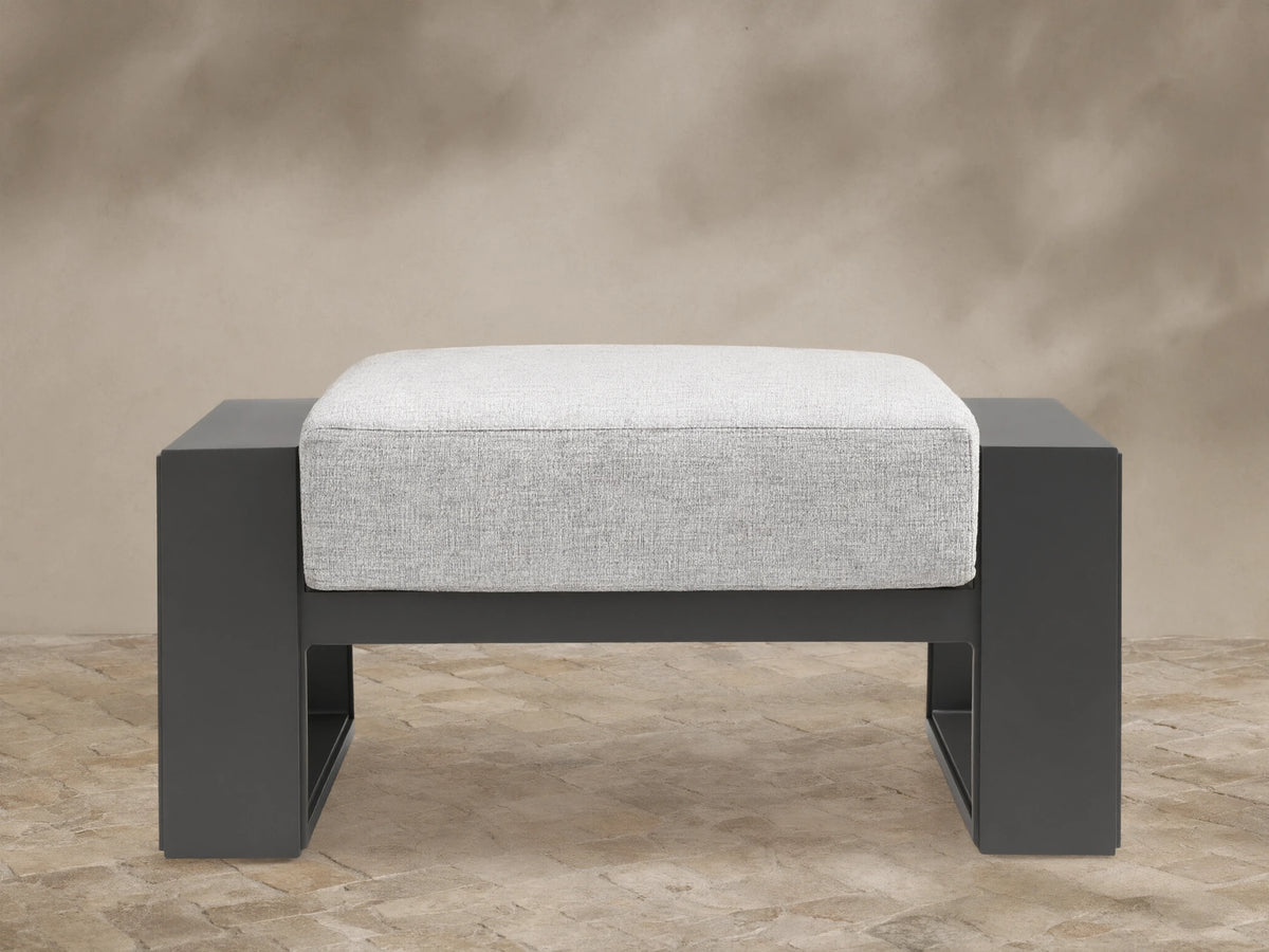 Canyon Outdoor Aluminum Ottoman with dark gray frame and light gray cushion, shown front-on on a tiled patio.