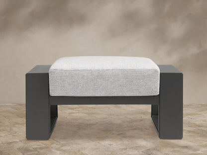 Canyon Outdoor Aluminum Ottoman with dark gray frame and light gray cushion, shown front-on on a tiled patio.