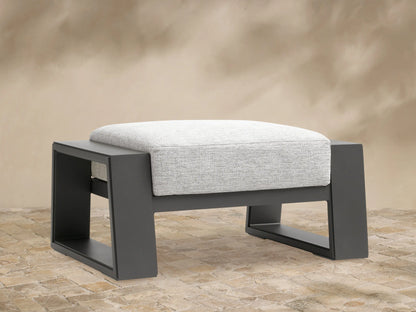 Canyon Outdoor Aluminum Ottoman with dark gray frame and light gray cushion, shown front-on on a tiled patio.