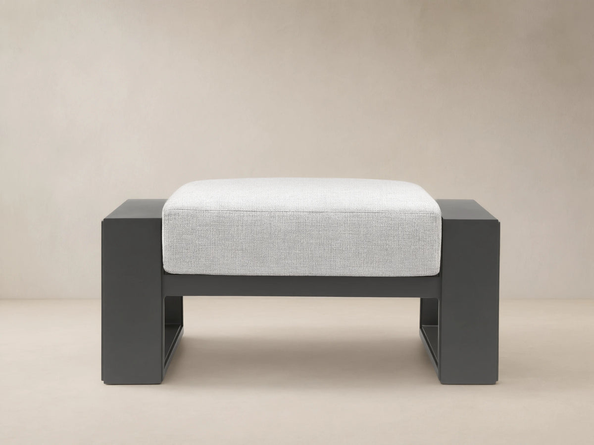 Canyon Outdoor Aluminum Ottoman with dark gray frame and light gray cushion, shown front-on on a tiled patio.