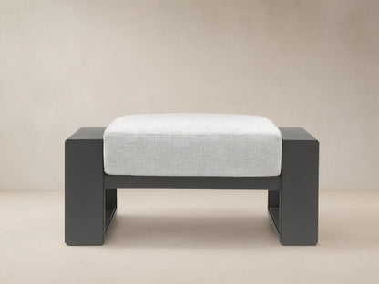 Canyon Outdoor Aluminum Ottoman with dark gray frame and light gray cushion, shown front-on on a tiled patio.