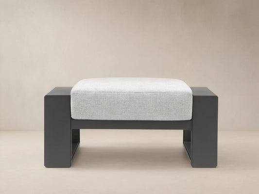 Canyon Outdoor Aluminum Ottoman with dark gray frame and light gray cushion, shown front-on on a tiled patio.