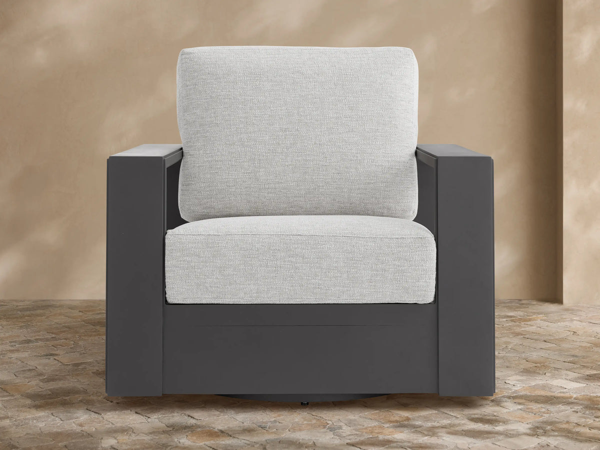 Canyon Outdoor Aluminum Swivel Chair, dark gray frame with light gray cushions, angled on a stone patio against a tan wall.