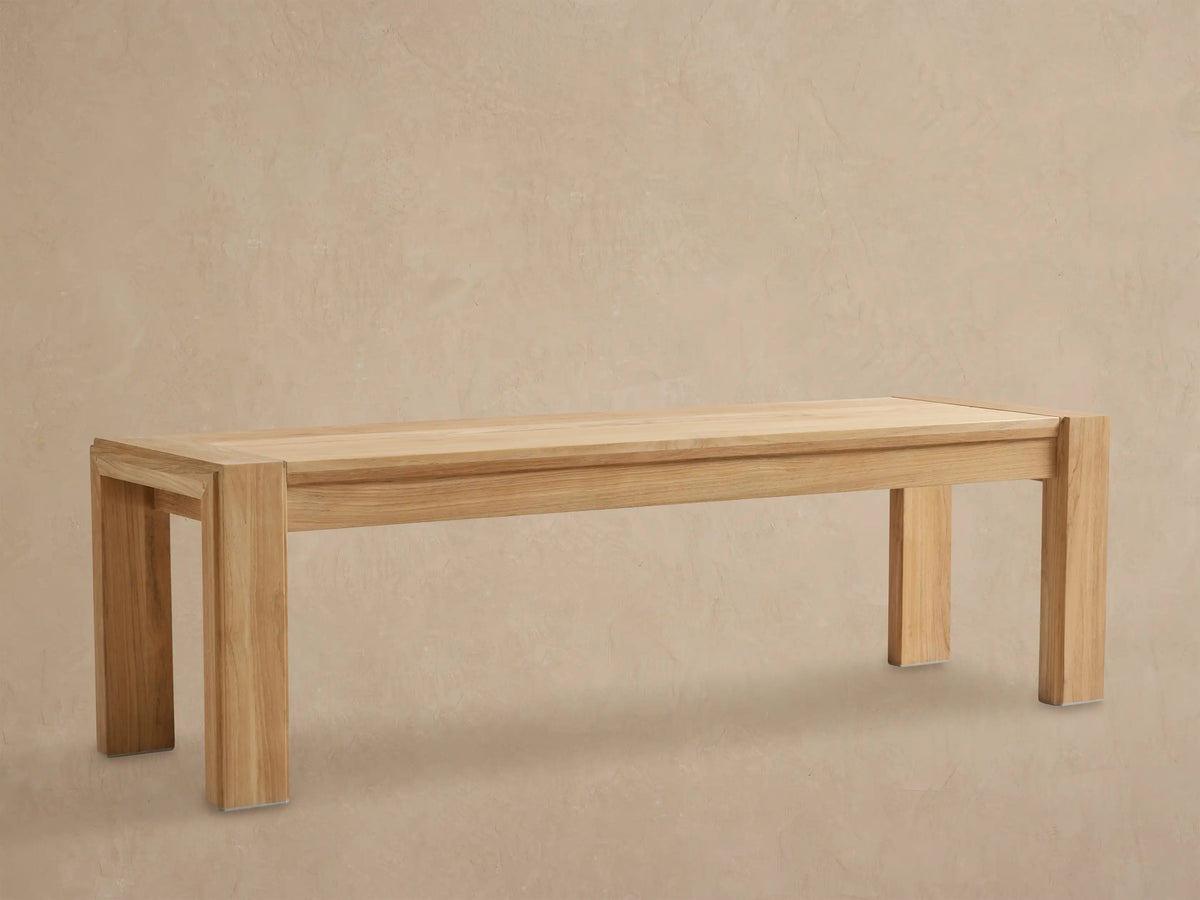 Canyon Outdoor Teak Dining Bench, light natural wood, rectangular with thick legs, centered on a stone floor against a textured wall.