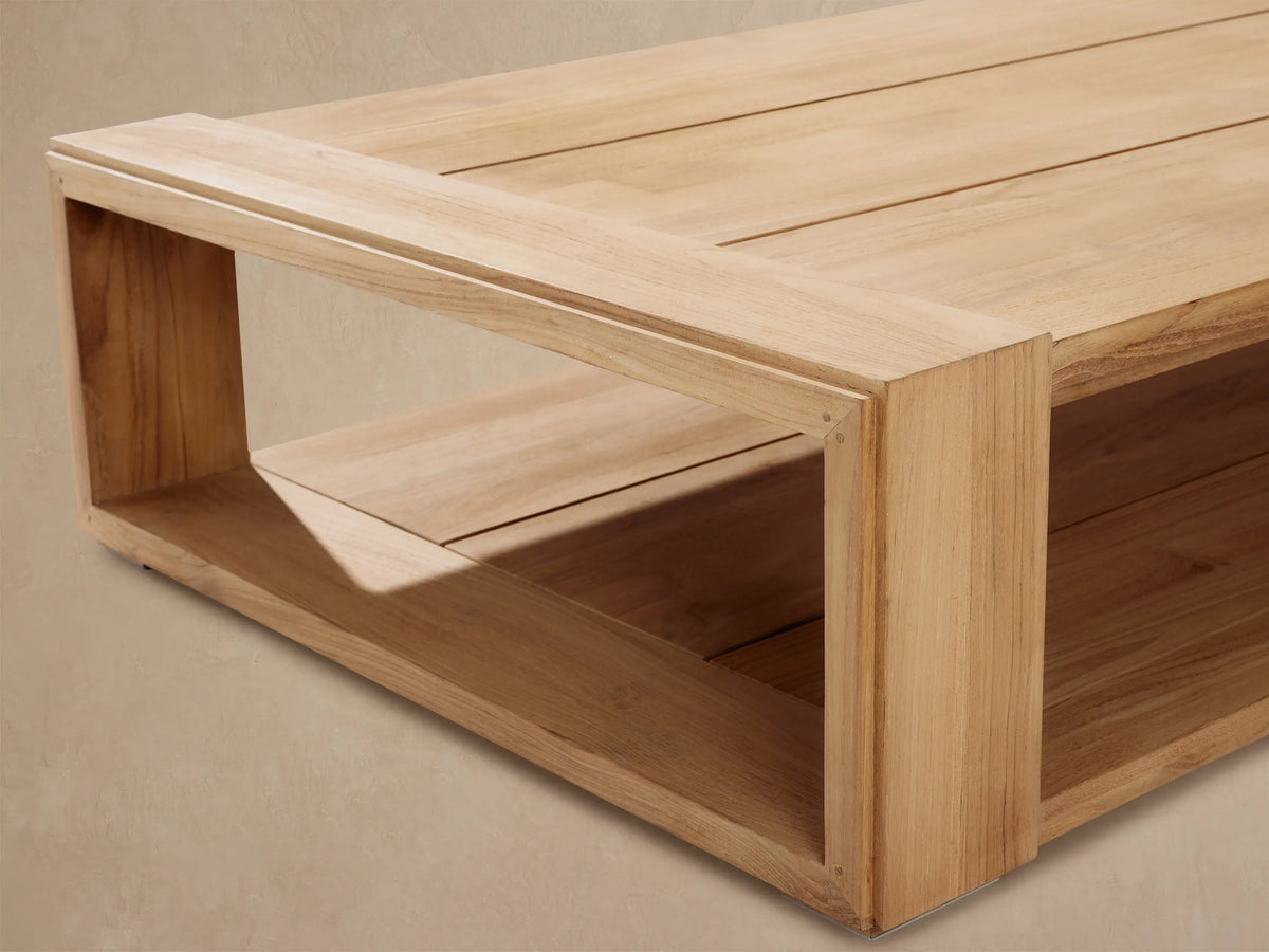 Canyon Outdoor Teak Coffee Table, rectangular light natural teak wood with slatted top and lower shelf on rustic patio.