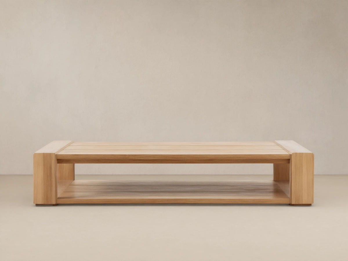Canyon Outdoor Teak Coffee Table: a rectangular light natural wood table with slatted top & lower shelf, on a stone patio.