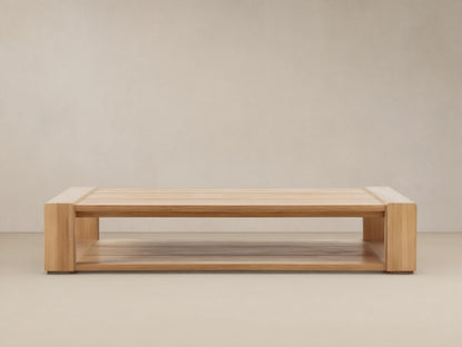 Canyon Outdoor Teak Coffee Table: a rectangular light natural wood table with slatted top & lower shelf, on a stone patio.