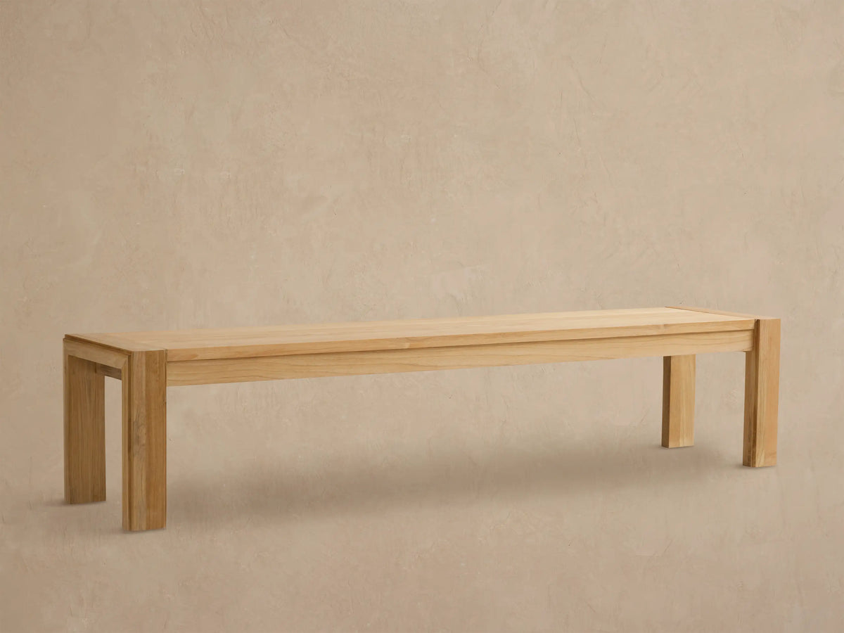 Canyon Outdoor Teak Dining Bench, light natural wood, rectangular with thick block legs, against a textured tan wall and stone floor.