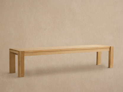Canyon Outdoor Teak Dining Bench, light natural wood, rectangular with thick block legs, against a textured tan wall and stone floor.