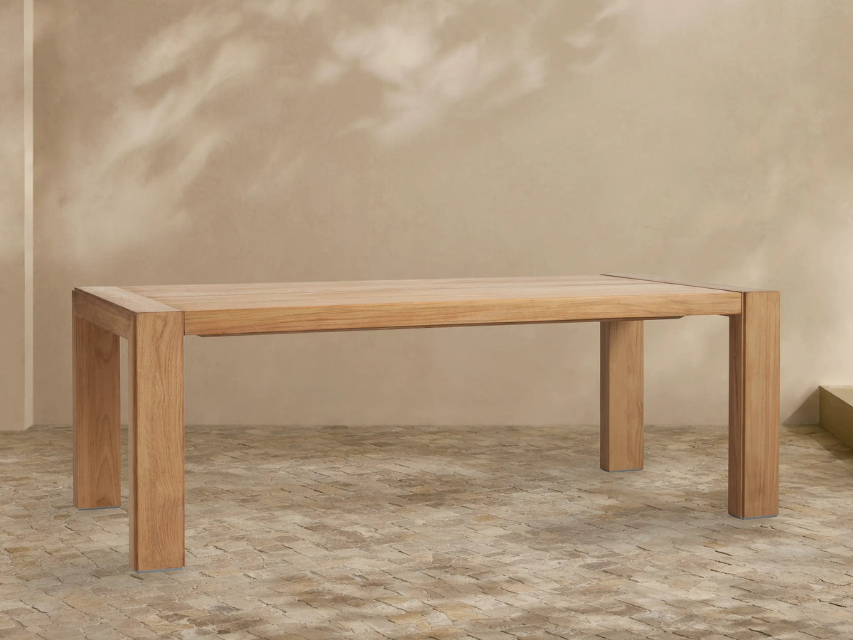 Canyon Outdoor Teak Dining Table, rectangular, natural teak wood. Frontal view in an outdoor setting with a light beige wall & tiled floor.