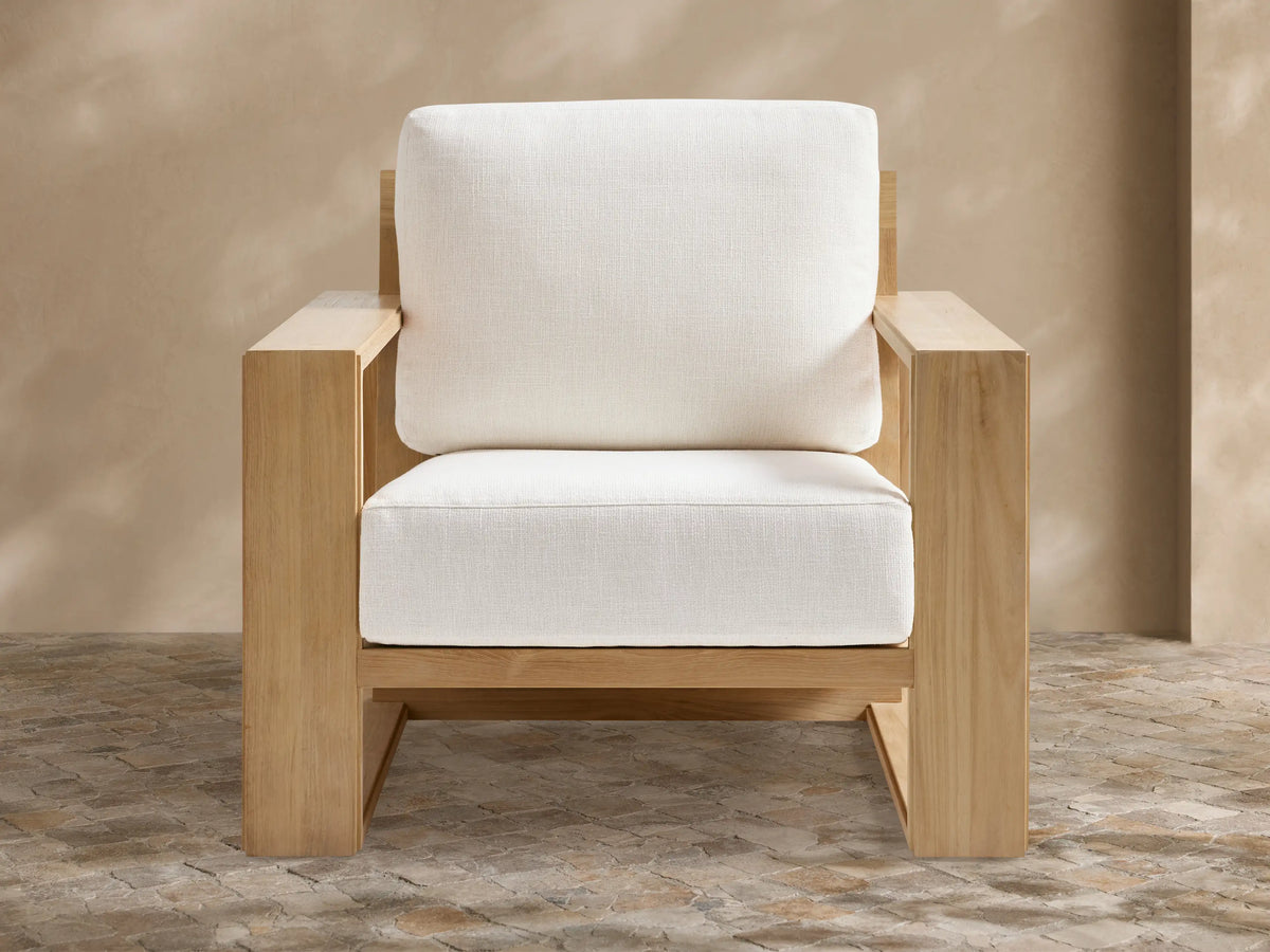 Canyon Outdoor Teak Lounge Chair. Modern, light-toned teak wood frame with thick white cushions, angled on a tiled patio.