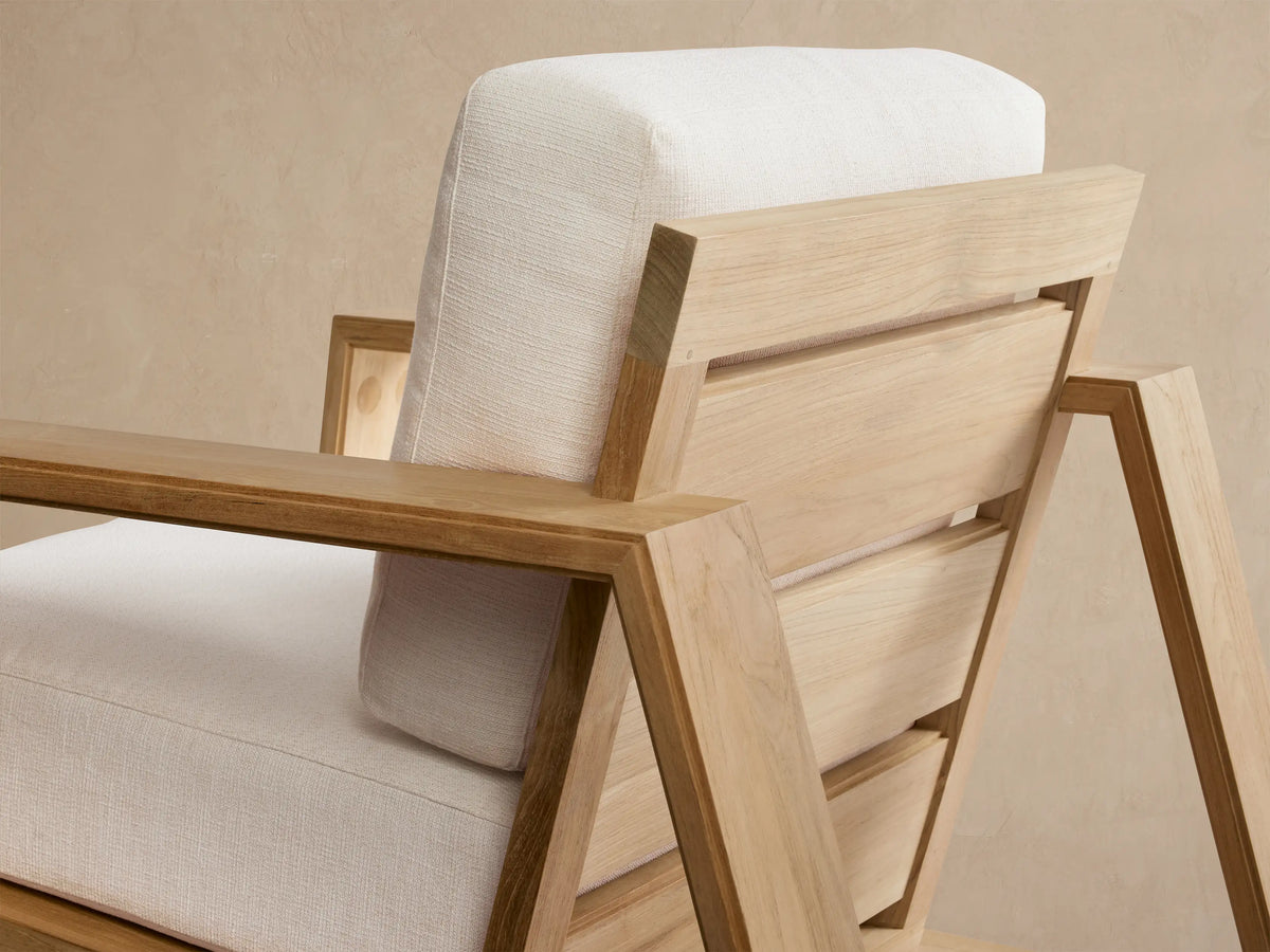 Front view of Canyon Outdoor Teak Lounge Chair: natural teak wood frame, thick white cushions, on a light stone patio.