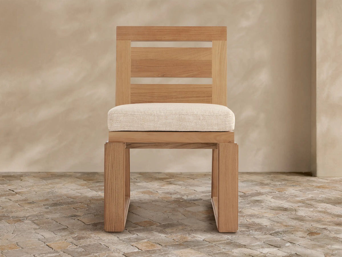 Light brown Canyon Teak Outdoor Dining Side Chair with cream cushion, modern slatted back & clear strips, angled on stone floor.