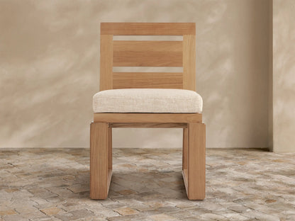 Light brown Canyon Teak Outdoor Dining Side Chair with cream cushion, modern slatted back & clear strips, angled on stone floor.