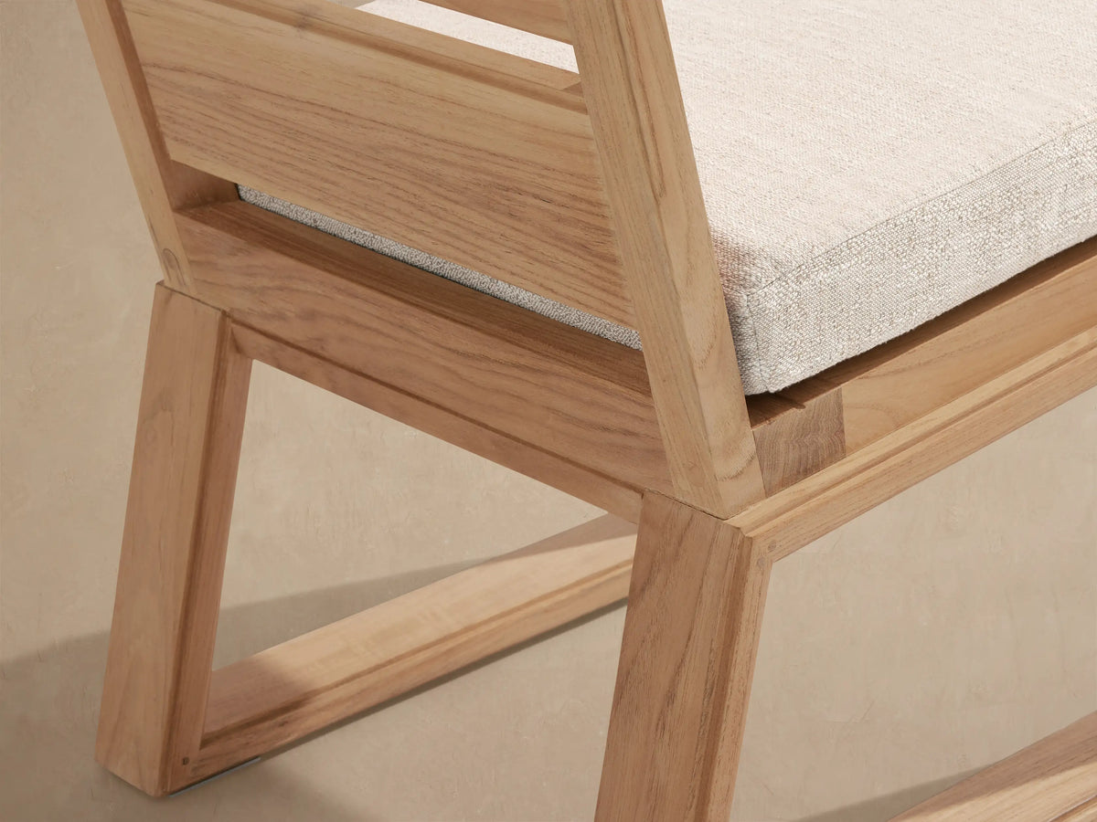 Canyon Outdoor Teak Dining Side Chair, front view. Natural teak wood frame, slatted back, and thick light beige seat cushion on tiled floor.