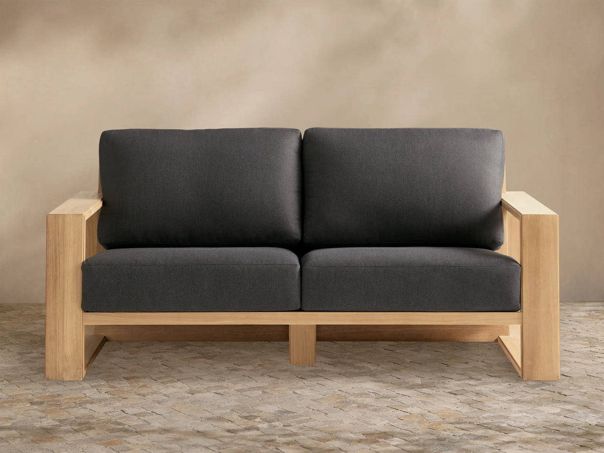 Canyon Outdoor Teak Sofa, a 2-seater with natural teak wood frame and dark gray cushions, on a stone patio against a beige wall.