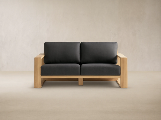 Canyon Outdoor Teak Sofa, a 2-seater with natural teak wood frame and dark gray cushions, on a stone patio against a beige wall.