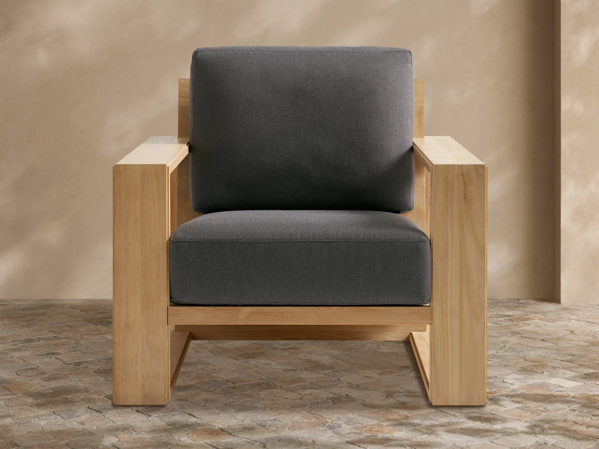 Canyon Outdoor Teak Lounge Chair with geometric natural teak wood frame and dark gray cushions on a stone patio.