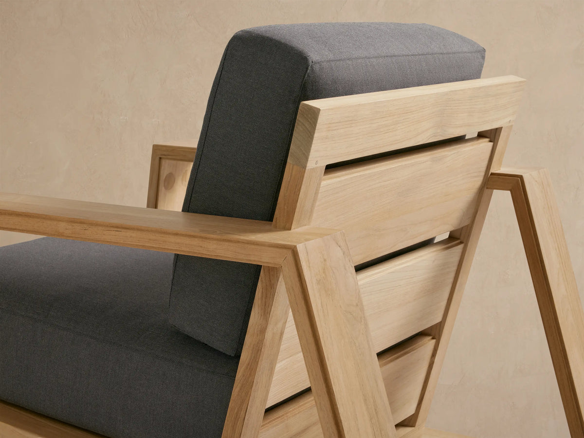 Canyon Outdoor Teak Lounge Chair, front view. Light natural teak frame with thick dark gray cushions on a tiled patio.